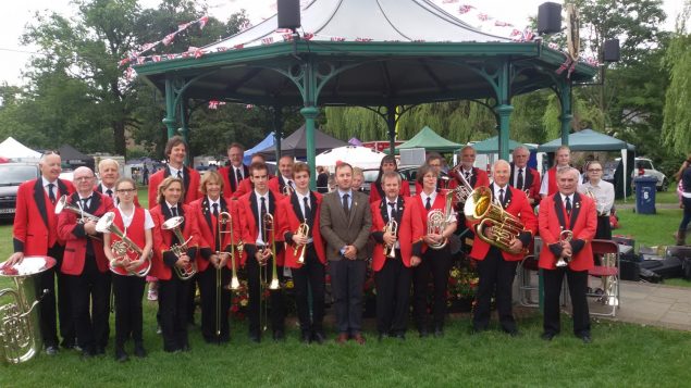 Group of male and female brass band members standing in front of band stand.