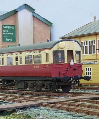 Red and cream train on a railway track, yellow building to the right, red and green building on the left.