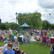 Groups of people, in the meadow, bandstand in background.