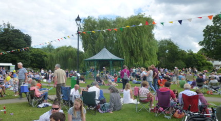 Groups of people, in the meadow, bandstand in background.