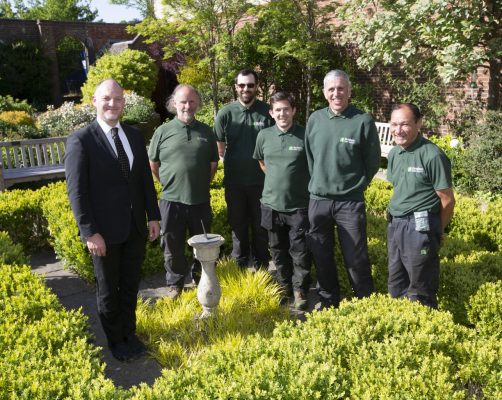 Man in suit with five men in green uniform standing in a garden