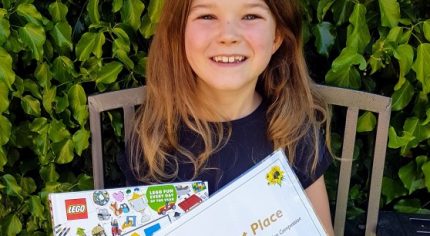 Girl sitting on bench in garden holding a certificate and lego prize.