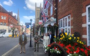 Armed Forces Day flag raising ceremony. Five people standing outside the town hall. Armed forces day flag and the Union flag are flying. Colourful flowers in a hop cart in the foreground.