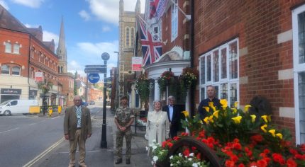 Armed Forces Day flag raising ceremony. Five people standing outside the town hall. Armed forces day flag and the Union flag are flying. Colourful flowers in a hop cart in the foreground.